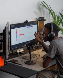 A person wearing headphones is sitting at a desk with a microphone, appearing to communicate or record a podcast. They are using a computer with a monitor displaying a web interface for creating an episode. Other desk items include a keyboard, a desk lamp, and some electronic devices. There's a small potted plant in the background.