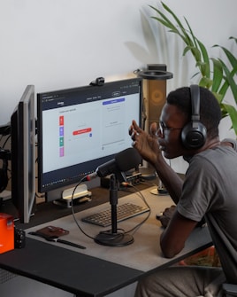 A person wearing headphones is sitting at a desk with a microphone, appearing to communicate or record a podcast. They are using a computer with a monitor displaying a web interface for creating an episode. Other desk items include a keyboard, a desk lamp, and some electronic devices. There's a small potted plant in the background.