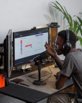 A person wearing headphones is sitting at a desk with a microphone, appearing to communicate or record a podcast. They are using a computer with a monitor displaying a web interface for creating an episode. Other desk items include a keyboard, a desk lamp, and some electronic devices. There's a small potted plant in the background.