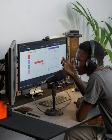A person wearing headphones is sitting at a desk with a microphone, appearing to communicate or record a podcast. They are using a computer with a monitor displaying a web interface for creating an episode. Other desk items include a keyboard, a desk lamp, and some electronic devices. There's a small potted plant in the background.