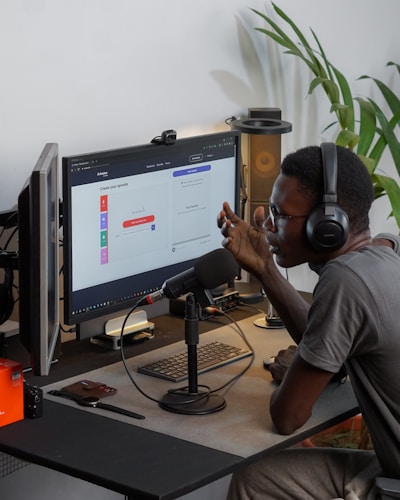 A person wearing headphones is sitting at a desk with a microphone, appearing to communicate or record a podcast. They are using a computer with a monitor displaying a web interface for creating an episode. Other desk items include a keyboard, a desk lamp, and some electronic devices. There's a small potted plant in the background.