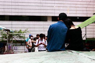 A couple is sitting closely together on a raised platform, with the woman resting her head on the man’s shoulder. The man is wearing a blue denim jacket, and the woman is dressed in black. In the background, a group of people is gathered, some of whom are wearing face masks and looking at their phones. The surrounding area includes a metal railing and an outdoor setting with some greenery.
