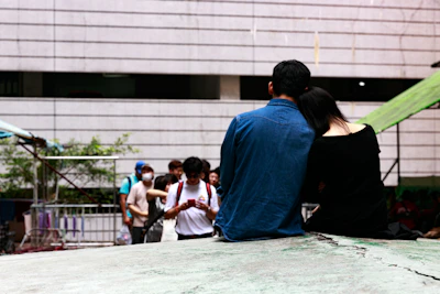 A couple is sitting closely together on a raised platform, with the woman resting her head on the man’s shoulder. The man is wearing a blue denim jacket, and the woman is dressed in black. In the background, a group of people is gathered, some of whom are wearing face masks and looking at their phones. The surrounding area includes a metal railing and an outdoor setting with some greenery.