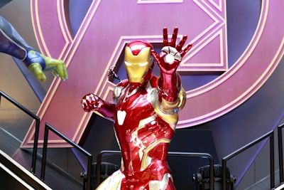 A life-sized figure of a superhero in red and gold armor is striking a dynamic pose with an outstretched hand. The background features a large, stylized letter 'A' in purple and pink. Part of another figure with a green hand and purple sleeve is visible, suggesting a scene of action or combat.