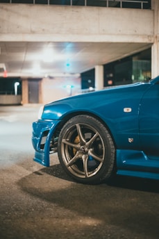 A close-up view of a blue sports car parked in a dimly lit garage. The focus is on the front wheel and bumper, which features a stylish design and alloy rims. The background is an urban setting, with concrete pillars and soft lighting providing a moody atmosphere.