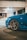 A close-up view of a blue sports car parked in a dimly lit garage. The focus is on the front wheel and bumper, which features a stylish design and alloy rims. The background is an urban setting, with concrete pillars and soft lighting providing a moody atmosphere.