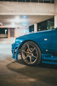 A close-up view of a blue sports car parked in a dimly lit garage. The focus is on the front wheel and bumper, which features a stylish design and alloy rims. The background is an urban setting, with concrete pillars and soft lighting providing a moody atmosphere.