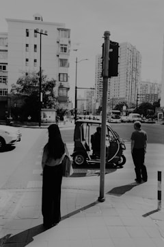 A cityscape with multi-story residential buildings in the background. In the foreground, a person stands with a bag, while another person is walking nearby. An auto rickshaw is parked on the street with a driver inside. A traffic light visible next to the road.
