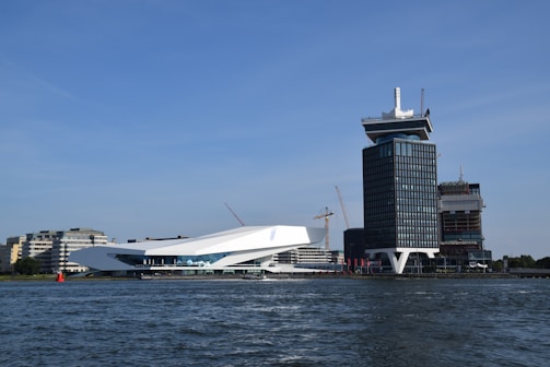 A waterfront scene featuring modern architecture with a sleek, white low-rise building alongside a taller, dark-colored tower. Cranes are visible in the background, indicating construction activity. The buildings stand next to a body of water under a clear blue sky.