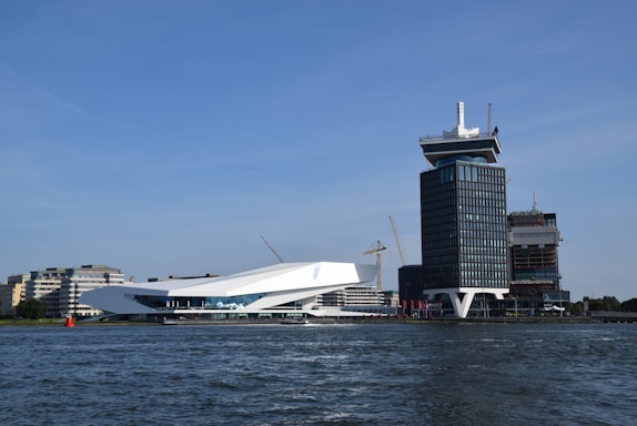 A waterfront scene featuring modern architecture with a sleek, white low-rise building alongside a taller, dark-colored tower. Cranes are visible in the background, indicating construction activity. The buildings stand next to a body of water under a clear blue sky.