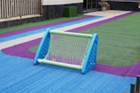 A small soccer goal sits on a colorful artificial turf field. The turf is marked with bright blue, green, and purple colors, creating a vibrant pattern. The goal is blue and green, and there is a ball visible inside it. The scene is likely part of a recreational or schoolyard area.