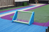 A small soccer goal sits on a colorful artificial turf field. The turf is marked with bright blue, green, and purple colors, creating a vibrant pattern. The goal is blue and green, and there is a ball visible inside it. The scene is likely part of a recreational or schoolyard area.