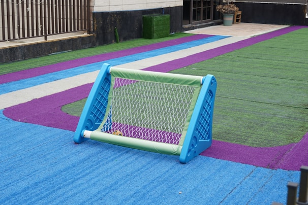 A small soccer goal sits on a colorful artificial turf field. The turf is marked with bright blue, green, and purple colors, creating a vibrant pattern. The goal is blue and green, and there is a ball visible inside it. The scene is likely part of a recreational or schoolyard area.