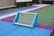 A small soccer goal sits on a colorful artificial turf field. The turf is marked with bright blue, green, and purple colors, creating a vibrant pattern. The goal is blue and green, and there is a ball visible inside it. The scene is likely part of a recreational or schoolyard area.