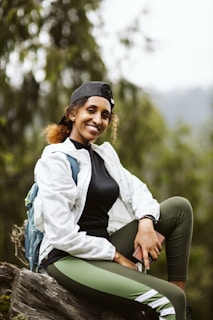 A person is sitting on a rock in an outdoor setting, wearing a white jacket, black shirt, green leggings, and a black cap. They are holding a smartphone and have a blue backpack. The background is a blurred natural forest environment.