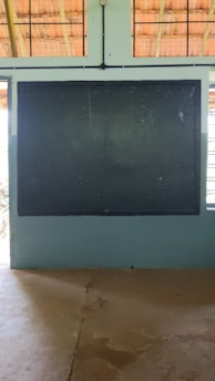 A traditional classroom setting with a large dark chalkboard mounted on a light blue wall. The ceiling has wooden beams and wire mesh, allowing natural light to filter through. The floor appears to be concrete.