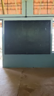 A traditional classroom setting with a large dark chalkboard mounted on a light blue wall. The ceiling has wooden beams and wire mesh, allowing natural light to filter through. The floor appears to be concrete.