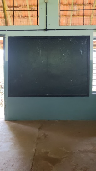 A traditional classroom setting with a large dark chalkboard mounted on a light blue wall. The ceiling has wooden beams and wire mesh, allowing natural light to filter through. The floor appears to be concrete.