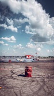 A car is drifting in a large open parking lot, creating smoke and skid marks on the asphalt. Around the area, red barrels and a cordoned-off section suggest a controlled environment for car stunts. In the background, a bright red fire truck is parked, under a partly cloudy sky.