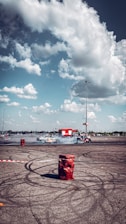 A car is drifting in a large open parking lot, creating smoke and skid marks on the asphalt. Around the area, red barrels and a cordoned-off section suggest a controlled environment for car stunts. In the background, a bright red fire truck is parked, under a partly cloudy sky.
