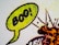 A close-up of a comic-style speech bubble with the word 'Boo!' in bold black letters inside. The bubble is colored with a bright green hue and has a textured pattern, typical of vintage comic book printing. The background features abstract lines and shapes with hints of orange and red colors.