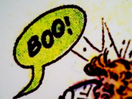 A close-up of a comic-style speech bubble with the word 'Boo!' in bold black letters inside. The bubble is colored with a bright green hue and has a textured pattern, typical of vintage comic book printing. The background features abstract lines and shapes with hints of orange and red colors.