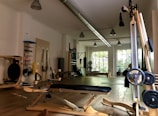 A well-lit gym space featuring various workout equipment including a wooden exercise bench, weightlifting bench, punching bag, and exercise balls. The room has large windows allowing natural light, wooden flooring, and equipment organized neatly along the walls.