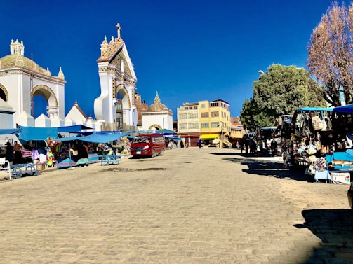A lively outdoor market scene in a cobblestone plaza, surrounded by historical architecture with ornate archways. Stalls with colorful fabrics and goods are covered by blue canopies with various vendors and shoppers interacting. A red vehicle is parked nearby, and there are large trees providing shade.