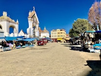 A lively outdoor market scene in a cobblestone plaza, surrounded by historical architecture with ornate archways. Stalls with colorful fabrics and goods are covered by blue canopies with various vendors and shoppers interacting. A red vehicle is parked nearby, and there are large trees providing shade.