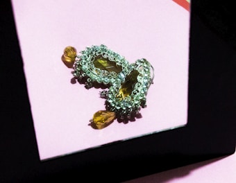 An ornate pair of earrings lies on a reflective surface. The earrings are designed with intricate patterns consisting of small crystals and larger teardrop-shaped amber stones. The reflection creates a sense of depth and echo in the composition. The background is a soft pink tone, while sharp contrasts are present due to strong lighting creating shadows.