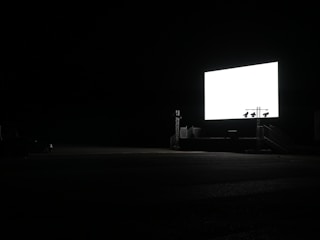 A large, illuminated screen stands in a dark, open space, likely a drive-in theater. The surroundings are mostly engulfed in darkness, with minimal visible structures.