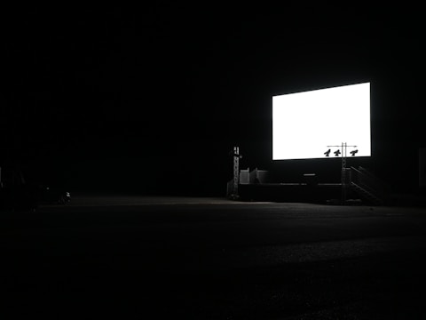 A large, illuminated screen stands in a dark, open space, likely a drive-in theater. The surroundings are mostly engulfed in darkness, with minimal visible structures.