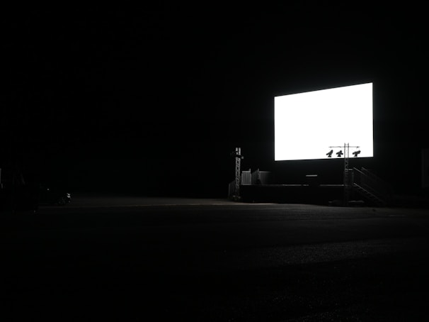 A large, illuminated screen stands in a dark, open space, likely a drive-in theater. The surroundings are mostly engulfed in darkness, with minimal visible structures.