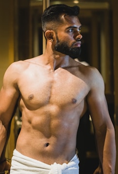 A man with a muscular build stands, partially covered with a white towel around his waist, in a dimly lit indoor setting. He has a beard and short hair, and is looking off to the side.