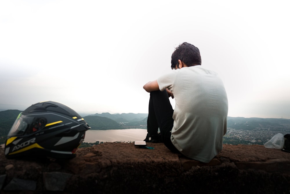 a man sitting on top of a cliff next to a motorcycle helmet