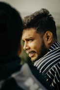 A person with facial hair is in profile view, wearing a striped shirt, and appears to be engaged in conversation with someone whose back is facing the camera.