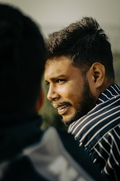 A person with facial hair is in profile view, wearing a striped shirt, and appears to be engaged in conversation with someone whose back is facing the camera.