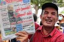 A man in a red shirt and black cap is holding a newspaper. The newspaper features an image of another man and various headlines in Arabic. The man holding the newspaper is smiling broadly. A person in the background wears a face mask, and there are trees visible in the distance.