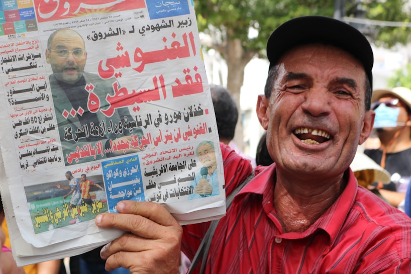 A man in a red shirt and black cap is holding a newspaper. The newspaper features an image of another man and various headlines in Arabic. The man holding the newspaper is smiling broadly. A person in the background wears a face mask, and there are trees visible in the distance.