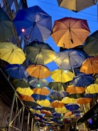 Multiple umbrellas in various colors are suspended in the air, creating a canopy effect above a street. Buildings flank the scene on either side, and there are lights interspersed among the umbrellas.