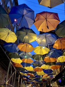 Multiple umbrellas in various colors are suspended in the air, creating a canopy effect above a street. Buildings flank the scene on either side, and there are lights interspersed among the umbrellas.