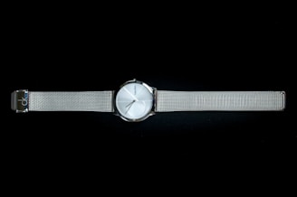 A sleek metallic wristwatch with a minimalist design, featuring a round silver dial and a slim mesh band. The watch face has simple hour markers and slender hands, with a brand name subtly printed on the dial. The watch band is adjustable and provides a modern, elegant look.