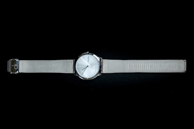 A sleek metallic wristwatch with a minimalist design, featuring a round silver dial and a slim mesh band. The watch face has simple hour markers and slender hands, with a brand name subtly printed on the dial. The watch band is adjustable and provides a modern, elegant look.