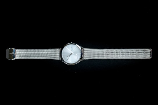 A sleek metallic wristwatch with a minimalist design, featuring a round silver dial and a slim mesh band. The watch face has simple hour markers and slender hands, with a brand name subtly printed on the dial. The watch band is adjustable and provides a modern, elegant look.