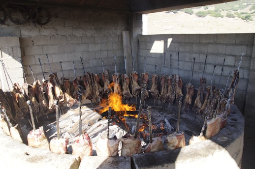 A circular barbecue setup with large sections of meat skewered and arranged around an open flame. The structure is made of stone or concrete, and the scene appears to be outdoors with a rustic vibe. The meat is being cooked over open fire, with the flames visible in the center.
