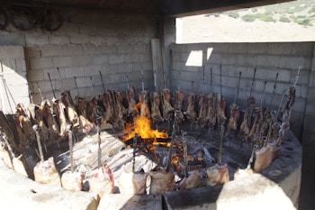 A circular barbecue setup with large sections of meat skewered and arranged around an open flame. The structure is made of stone or concrete, and the scene appears to be outdoors with a rustic vibe. The meat is being cooked over open fire, with the flames visible in the center.