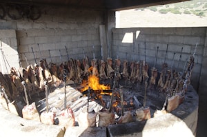 A circular barbecue setup with large sections of meat skewered and arranged around an open flame. The structure is made of stone or concrete, and the scene appears to be outdoors with a rustic vibe. The meat is being cooked over open fire, with the flames visible in the center.