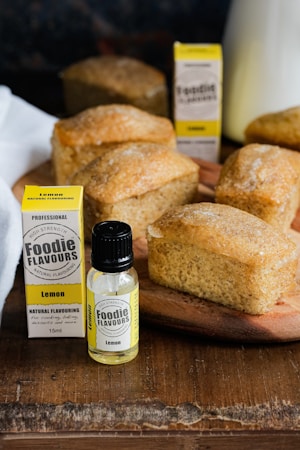 Several small, rectangular lemon cakes are presented on a wooden surface alongside bottles and boxes of lemon natural flavouring. The cakes have a light brown crust and look freshly baked. The flavouring bottles have a black cap and yellow labeling.