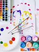 A collection of art supplies is displayed, including a watercolor palette with various colors, brushes, and several containers of vibrant paints. There's a paper plate used as a palette with dabs of paint, along with sheets of paper featuring simple, colorful paintings of two people. The scene is bright and colorful, suggesting an art or craft activity.