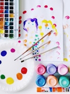 A collection of art supplies is displayed, including a watercolor palette with various colors, brushes, and several containers of vibrant paints. There's a paper plate used as a palette with dabs of paint, along with sheets of paper featuring simple, colorful paintings of two people. The scene is bright and colorful, suggesting an art or craft activity.